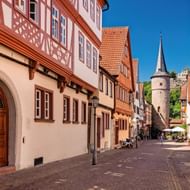 Smalle geplaveide straat in Karlstadt met traditionele vakwerkhuizen. Een middeleeuwse stenen toren met spitse torenspits rijst op de achtergrond.