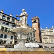 Witte marmeren fontein met klassiek standbeeld op Piazza delle Erbe in Verona, omringd door historische gebouwen met balkons en rode bakstenen toren.
