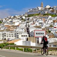 Fietser met rugzak bij plaatsnaambord van Baena langs de weg, met witte heuveldorp en kerktorens op de achtergrond.
