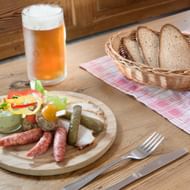 Houten bord met Duitse worsten, augurken en salade naast een glas bier en mand met donker brood op geruit tafelkleed.