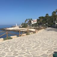 Kustpromenade in Denia, Costa Blanca Stenen kustpromenade in Denia met houten leuningen uitkijkend over de Middellandse Zee, palmbomen, witte gebouwen en vuurtoren zichtbaar.