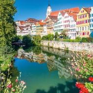 Rij kleurrijke vakwerkhuizen langs de Main in Tübingen met kerktoren, weerspiegeld in kalm water. Bloemen bloeien op voorgrond.