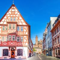 Geplaveide straat in de oude binnenstad van Mainz met kleurrijke vakwerkhuizen en barokke domtorens zichtbaar op de achtergrond.