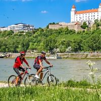 Twee fietsers in rode shirts met fietsen op grasrijke Donauoever. Kasteel van Bratislava met witte muren en rood dak zichtbaar op heuvel.