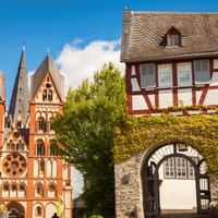 Rood-witte Dom van Limburg met drie torens naast een traditioneel vakwerkhuis met stenen boog, omringd door groene bomen onder blauwe lucht.