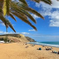 Zandstrand in Lagos, Algarve met palmtakken erboven, gouden kliffen op de achtergrond, strandgangers en tenten op het zand, blauwe zeegolven.