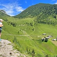 Vrouwelijke wandelaar met blauwe rugzak kijkt uit over de groene Rinnbergalm bergweidedal met traditionele alpenhutten en beboste bergen.