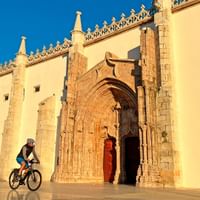 Fietsers bij historische kathedraal in Portugal Twee fietsers met helmen rijden langs een gotische kathedraal met sierlijke stenen ingang en boogvormige deur onder heldere blauwe hemel.
