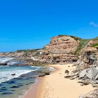 Portugese kustrotsen en zandstrand Rotsachtige kustlijn met gelaagde zandsteenrotsen, zandstrand en turquoise oceaangolven. Witte gebouwen zichtbaar op rotsen onder blauwe lucht.
