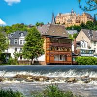 Marburg stadsgezicht met historisch kasteel op heuvel, kleurrijke vakwerkhuizen langs rivieroever en Lahn rivier met stuw op voorgrond.
