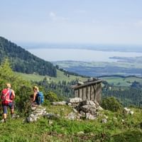 Twee wandelaars met rugzakken op een bergpad met uitzicht op de Chiemsee. Een houten schuilplaats op rotsen, beboste toppen en dal beneden.