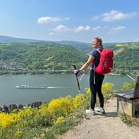 Wandelaarster met rode rugzak en wandelstokken staat op uitkijkpunt met uitzicht op de Rijn met cruiseschip en groene heuvels.