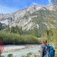Twee wandelaars met rugzakken bij een turquoise rivier, kijkend naar dramatische Alpentoppen van de Zugspitze-regio.