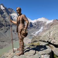 Houten wandelaar standbeeld met wandelstok op rotsuitsteeksel met uitzicht op Grossglockner gletsjer en besneeuwde bergen in Oostenrijk onder blauwe lucht.