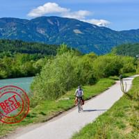 Fietser op verharde weg langs turquoise rivier met groene bergen op achtergrond. Rode bestseller stempel zichtbaar in hoek.