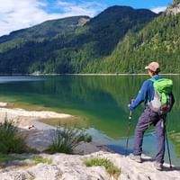 Wandelaar met groene rugzak en wandelstokken staat op rotsachtige oever van Schwarzensee meer, omringd door beboste bergen in Oostenrijk.