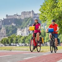Twee fietsers met fietstassen op rood fietspad langs de Salzach in Salzburg met vesting Hohensalzburg op de heuvel.