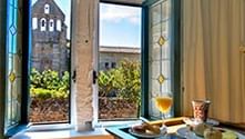 Hotelkamer interieur met open openslaande deuren die een historische stenen toren en tuin tonen. Een ontbijtblad met drankjes en eten staat op een tafel.
