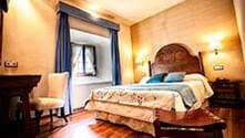 Warme hotelkamer met gesneden houten hoofdeinde, blauwe kussens, hardhouten vloer, open haard en houten kist aan voeteneind.