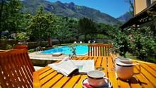 Houten terras met koffiekopjes en krant met uitzicht op een blauw zwembad, omgeven door weelderige tuinen en Picos de Europa bergen.