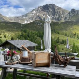 Houten terrastafel bij Mahdalm met traditionele Alpenversiering, bloemen en houten voorwerpen. Berghut en markante toppen op achtergrond.