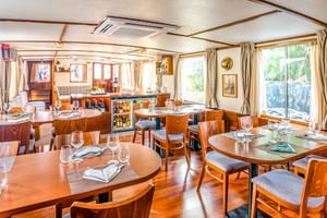 Lichte eetzaal op MS Caprice met houten tafels gedekt met glazen en servetten, gestoffeerde stoelen, grote ramen met gordijnen en een bar.