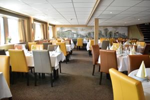 Interieur van MS De Willemstad scheepsrestaurant met kleurrijke gestoffeerde stoelen, witte tafelkleden en historische Nederlandse grachten muurschilderingen.