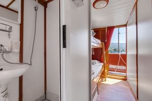 Compacte badkamer op MS Mirabela met douche, wastafel en spiegel. Zicht door open deur toont hut met bedden en raam met uitzicht op Kroatische kust.