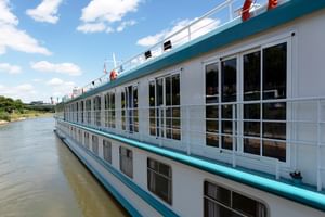 Wit MS Switzerland riviercruiseschip met blauwe rand en grote ramen, afgemeerd aan een kalme rivier onder blauwe lucht.