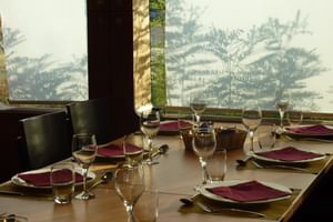 Restauranttafel gedekt met bordeauxrode servetten, wijnglazen en couverts aan boord van MS Ave Maria. Grote ramen tonen tropische vegetatie.