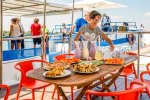 Bemanningslid serveert eten en drinken op het zonnedek van MS Caprice. Tafel met gebak, fruit en drankjes. Passagiers ontspannen op blauw-wit bootdek.