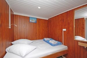 Compacte MS Kazimir scheepshut met houten wanden, wit plafond, eenpersoonsbed met wit beddengoed, blauwe handdoeken, leeslamp en spiegel.