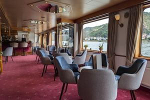 Modern lounge-interieur op MS Veloria met grijze fauteuils, bordeauxrood tapijt en grote ramen met uitzicht op rivier en groene heuvels.