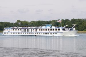 Wit riviercruiseschip MS De Willemstad varend op kalm water met beboste kustlijn op de achtergrond onder bewolkte hemel.