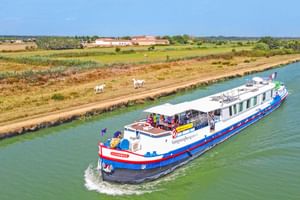 MS Caprice riviercruiseschip met blauwe en rode strepen varend op een kanaal. Landelijk landschap met velden en witte paarden op de oever.