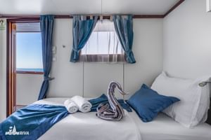 Lichte hut op MY Olmissum met wit bed, blauwe gordijnen, raam en deur met zeezicht. Handdoeken en decoratieve zwaan op bed.