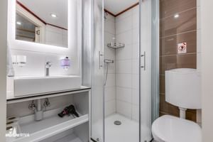 Compacte witte badkamer op MY Olmissum met wastafel, spiegel, glazen douchecabine met gordijn en toilet. Moderne voorzieningen met houteffect tegels.