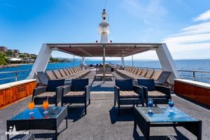 Zonnedek van cruiseschip MY Vita met rijen ligstoelen onder een luifel, rieten zitjes met blauwe drankjes en oceaanzicht onder heldere hemel.