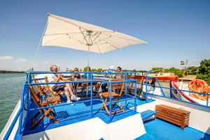 Zonnedek van MS Caprice met blauwe reling, witte parasol, houten ligstoelen en ontspannende passagiers. Rivier en boten op de achtergrond.