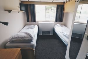 Compacte tweepersoonshut met twee eenpersoonsbedden, wit beddengoed, blauwe gordijnen, raamzicht en donker tapijt aan boord van De Willemstad.