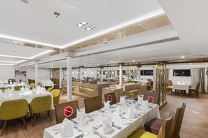 Modern restaurant interieur met wit gedekte tafels, gele en bruine stoelen, witte pilaren en houten vloer. Meerdere zitgedeeltes zichtbaar.