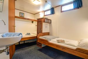 Compacte cabine op MS Flora met twee eenpersoonsbedden met houten frames, wastafel, kast en twee kleine ramen met blauwe gordijnen.