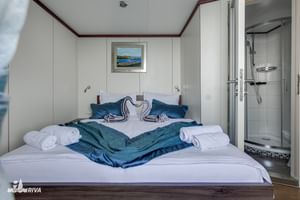 Lichte hut op MY Olmissum met tweepersoonsbed, wit beddengoed, blauwe handdoeken, kustfoto en glazen douchecabine.