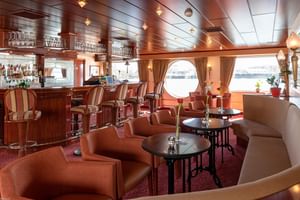 Elegante salon aan boord van MS Switzerland met houten bar, oranje leren stoelen, ronde tafels en rond patrijspoort met uitzicht op water.