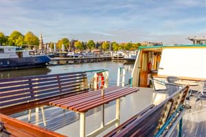 Zonnig dek van MS Flora met houten banken, witte reling en reddingsboei. Jachthaven met boten en bomen op achtergrond onder blauwe lucht.