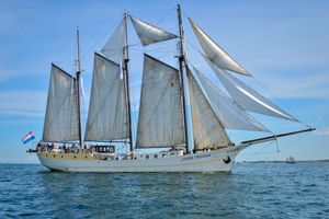 Driemast zeilschip Mare Frisium met witte zeilen op kalm blauw water onder heldere hemel, Nederlandse vlag zichtbaar.