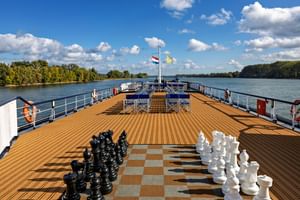 Zonnedek van MS Veloria met teakhouten vloer, reuzenschaakbord, blauwe ligstoelen en riviergezicht met groene oevers onder blauwe lucht.