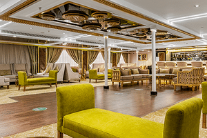 Ruime Nebu lounge met limoengroene fauteuils en banken, witte zuilen, versierd plafond en beige gordijnen. Donkere houten vloer en bar zichtbaar.