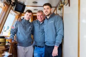 Drie bemanningsleden in bijpassende blauwe jassen staan samen in de houten binnencabine van het MY Vita schip.