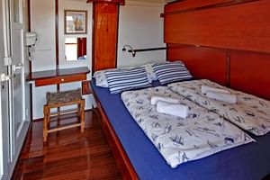 Scheepshut interieur met tweepersoonsbed met blauwe matras en nautisch beddengoed. Houten lambrisering, klein bureau met stoel en patrijspoort zichtbaar.